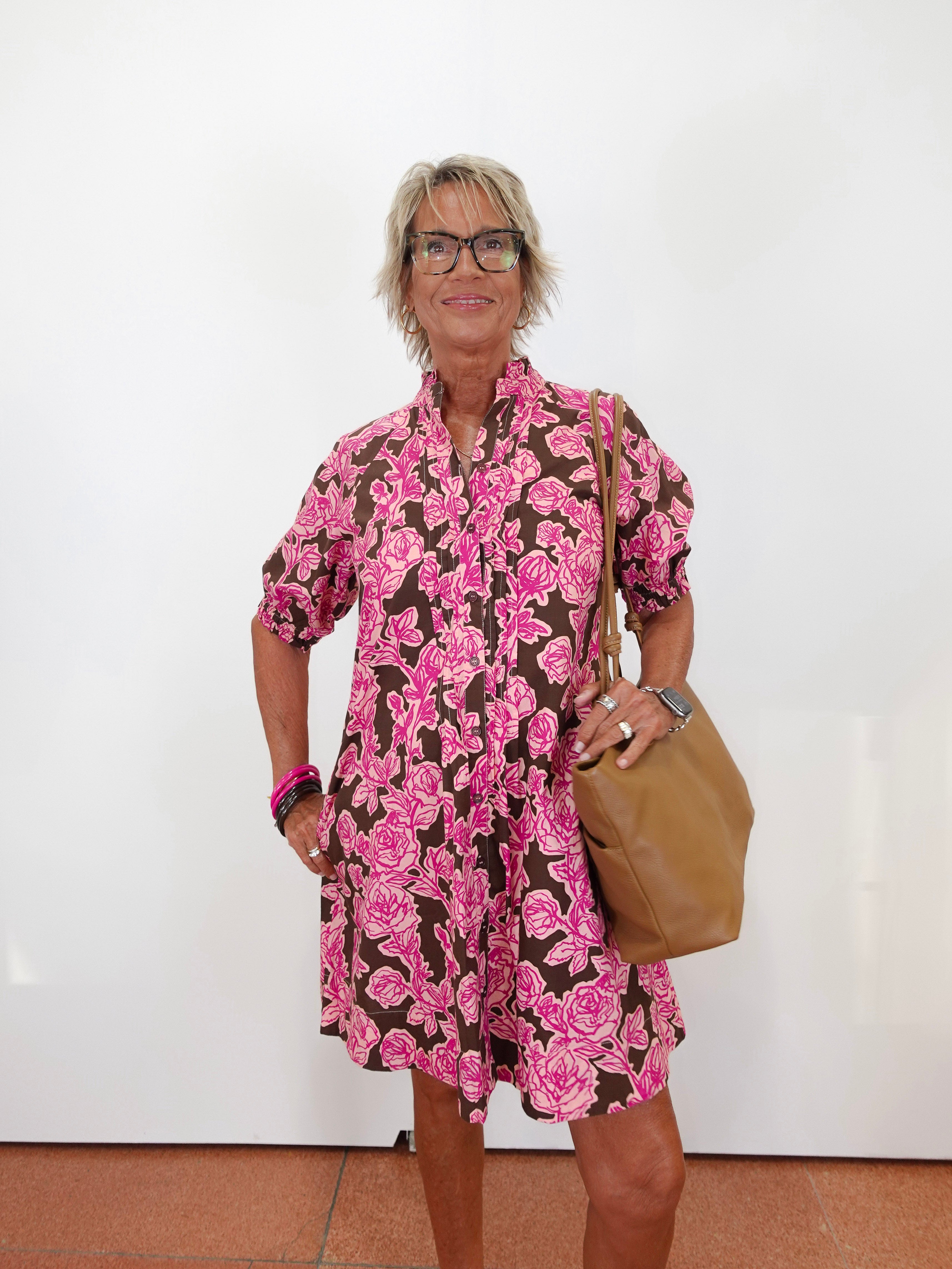 A woman with short blonde hair and glasses models a brown and pink floral print mini dress, holding a tan leather shoulder bag against a plain white wall.