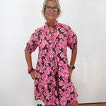 A woman with short blonde hair and glasses wearing a brown and pink floral print dress, standing against a plain white wall.