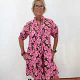A woman with short blonde hair and glasses wearing a brown and pink floral print dress, standing against a plain white wall.