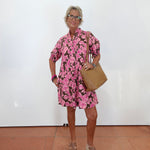 An older woman with short gray hair and glasses stands against a white wall wearing a pink and black floral print mini dress, a tan shoulder bag, and beige platform sandals.