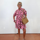 An older woman with short gray hair and glasses stands against a white wall wearing a pink and black floral print mini dress, a tan shoulder bag, and beige platform sandals.