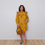 Woman wearing a mustard yellow dress with floral patterns against a white wall.