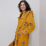 Woman wearing a yellow coat with floral embroidery against a white wall