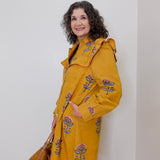 Woman wearing a yellow coat with floral embroidery against a white wall