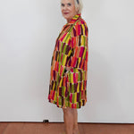 A woman with short white hair and a blue headband stands in profile wearing a colorful geometric print tunic dress with vertical blocks of red, yellow, green, and brown on a white wall and terracotta floor.