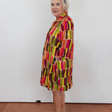 A woman with short white hair and a blue headband stands in profile wearing a colorful geometric print tunic dress with vertical blocks of red, yellow, green, and brown on a white wall and terracotta floor.