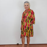 An older woman stands against a plain white wall wearing a short-sleeve collared dress with a bold geometric pattern in red, yellow, green, and brown, paired with tan wedge sandals.