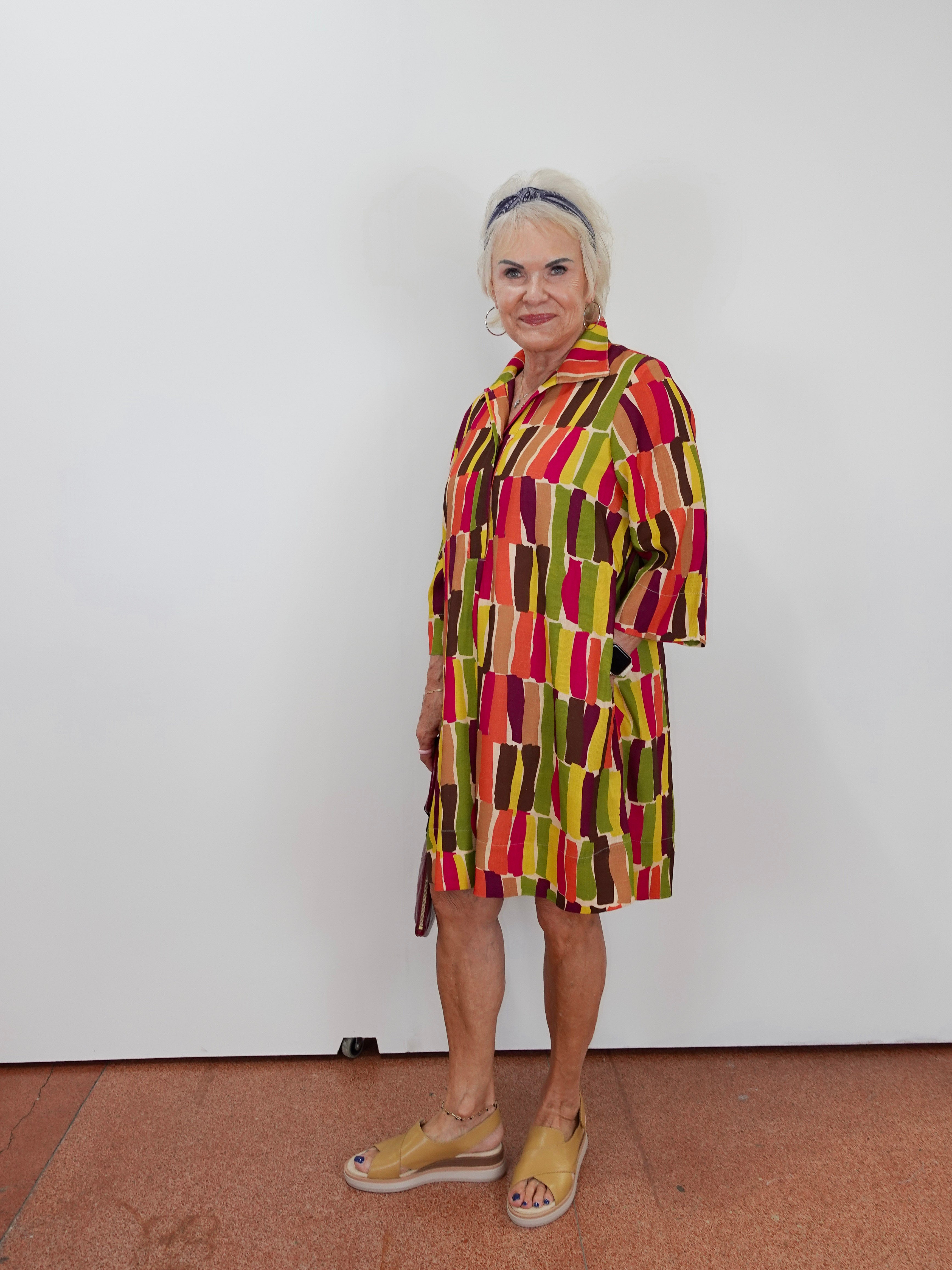 An older woman stands against a plain white wall wearing a short-sleeve collared dress with a bold geometric pattern in red, yellow, green, and brown, paired with tan wedge sandals.