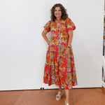 Woman wearing a colorful floral dress standing against a white wall.