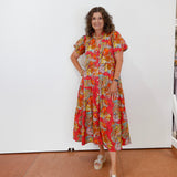 Woman wearing a colorful floral dress standing against a white wall.