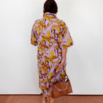Woman wearing a patterned dress holding a brown bag against a white wall.