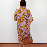 Woman wearing a patterned dress holding a brown bag against a white wall.
