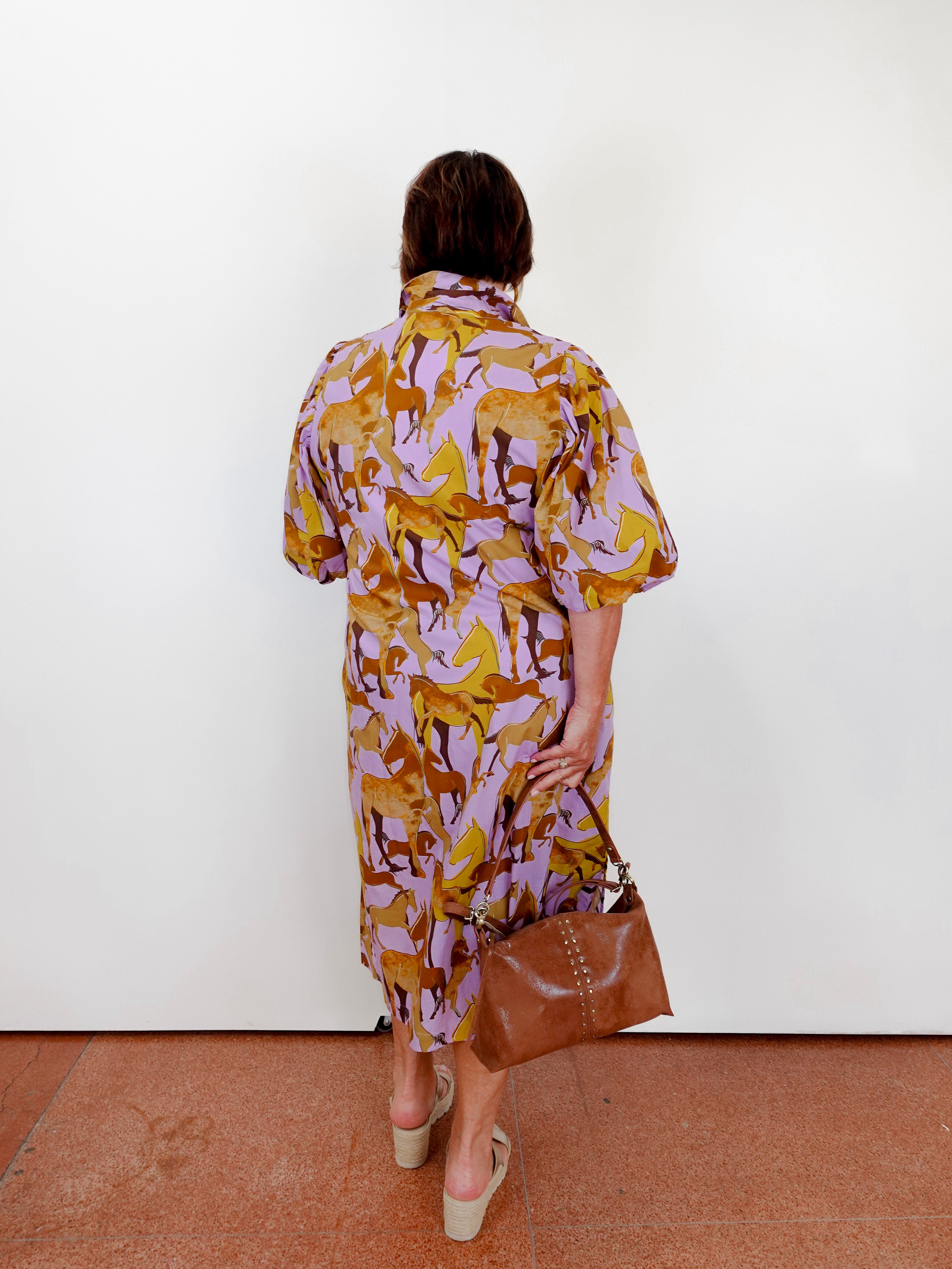 Woman wearing a patterned dress holding a brown bag against a white wall.