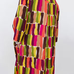 A long-sleeve button-up shirt with a colorful abstract geometric pattern of vertical brushstroke blocks in pink, orange, yellow, green, brown, and beige, shown from the back on a mannequin against a plain white wall.