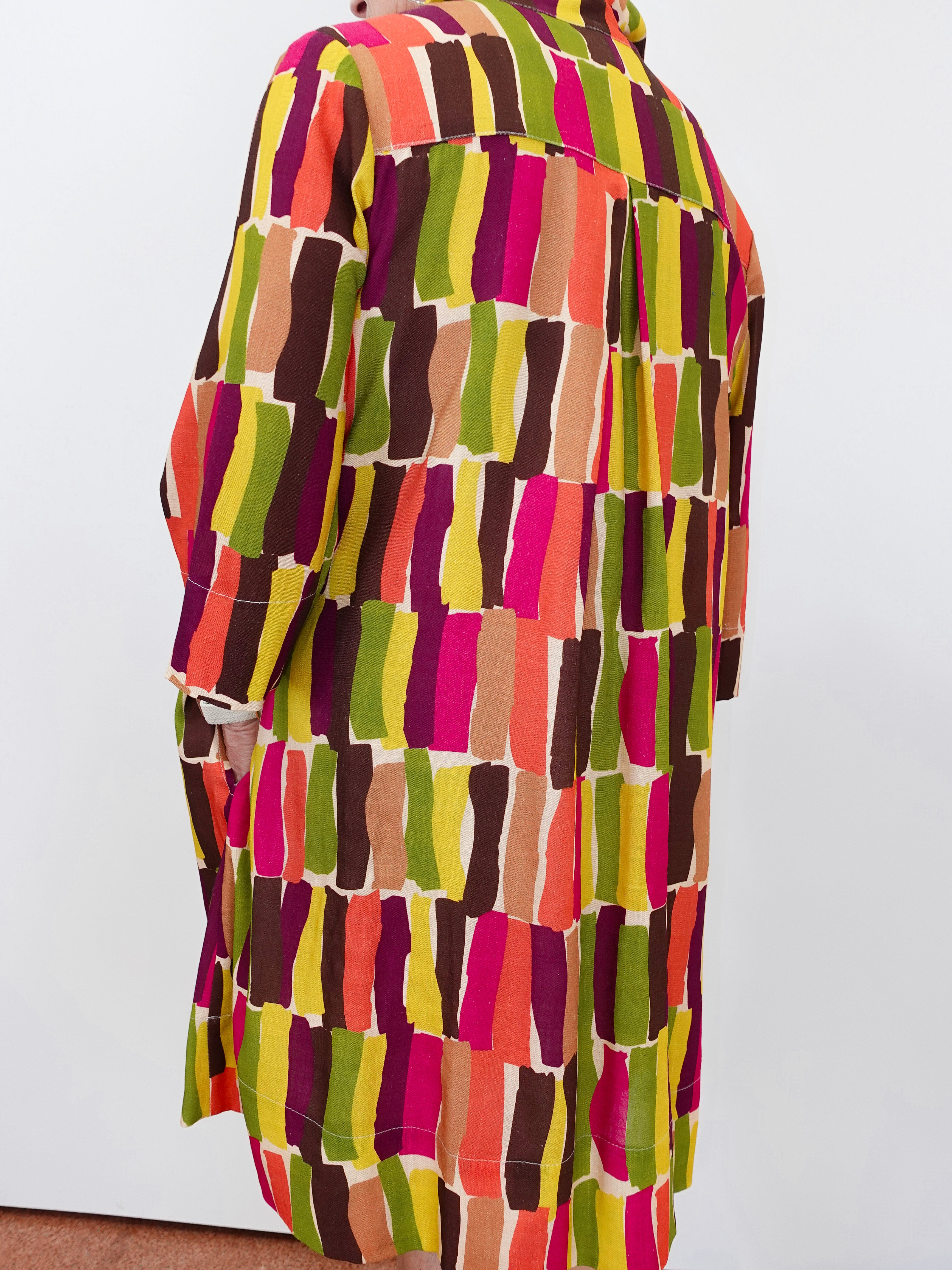 A long-sleeve button-up shirt with a colorful abstract geometric pattern of vertical brushstroke blocks in pink, orange, yellow, green, brown, and beige, shown from the back on a mannequin against a plain white wall.