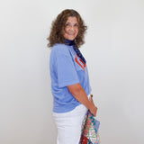 Woman wearing a blue shirt and white pants with a colorful scarf, standing against a plain white wall.
