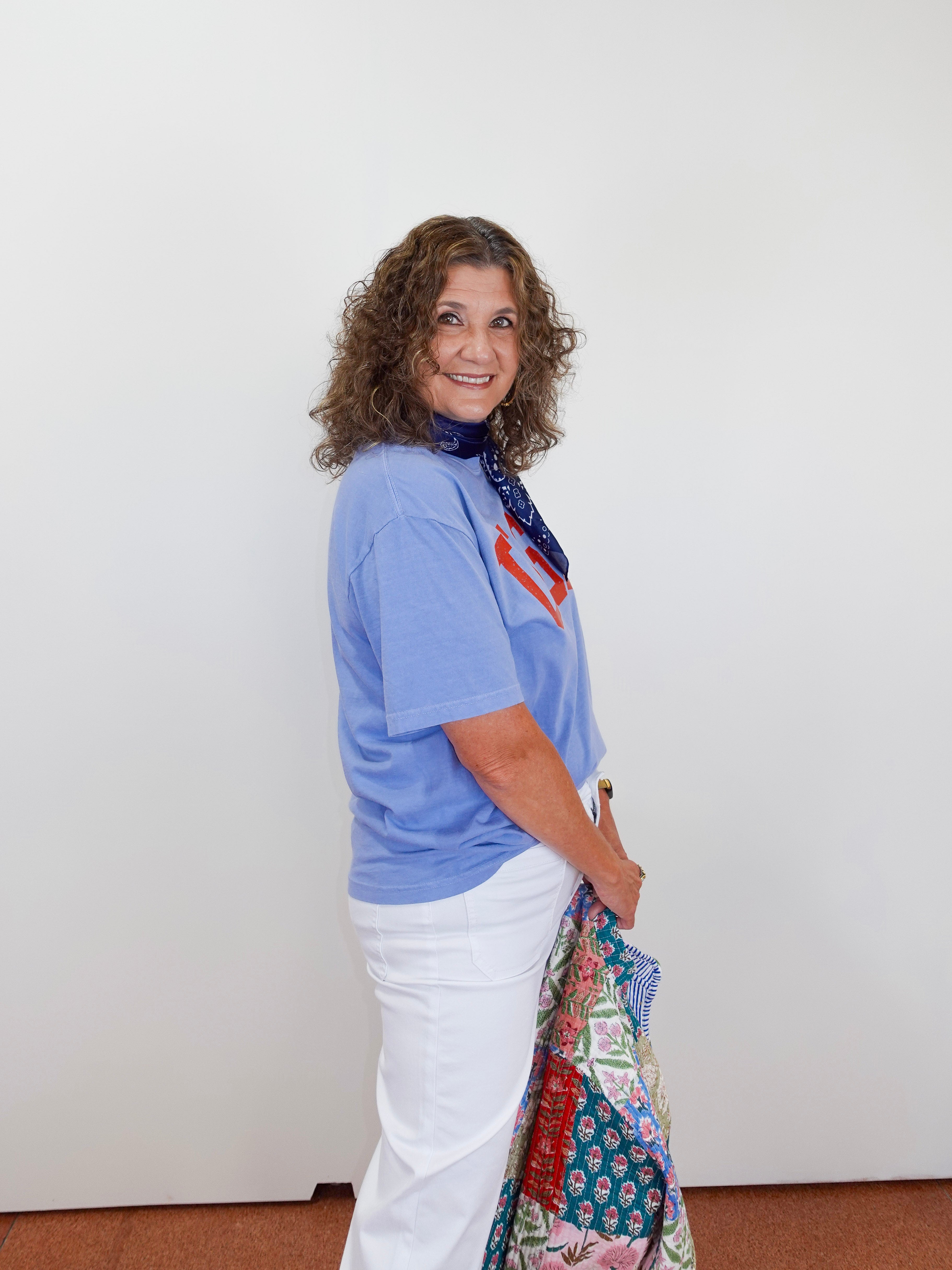 Woman wearing a blue shirt and white pants with a colorful scarf, standing against a plain white wall.