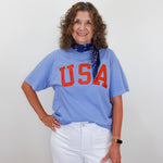 Woman wearing a blue 'USA' t-shirt with white pants against a plain background