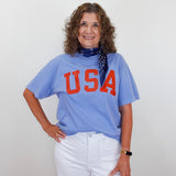 Woman wearing a blue 'USA' t-shirt with white pants against a plain background