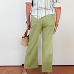 Person wearing a striped shirt and green pants with a beige handbag against a white wall.