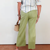 Person wearing a striped shirt and green pants with a beige handbag against a white wall.