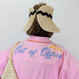 Person wearing a pink shirt with 'Out of Office' text, beige visor, and straw bag on a white background