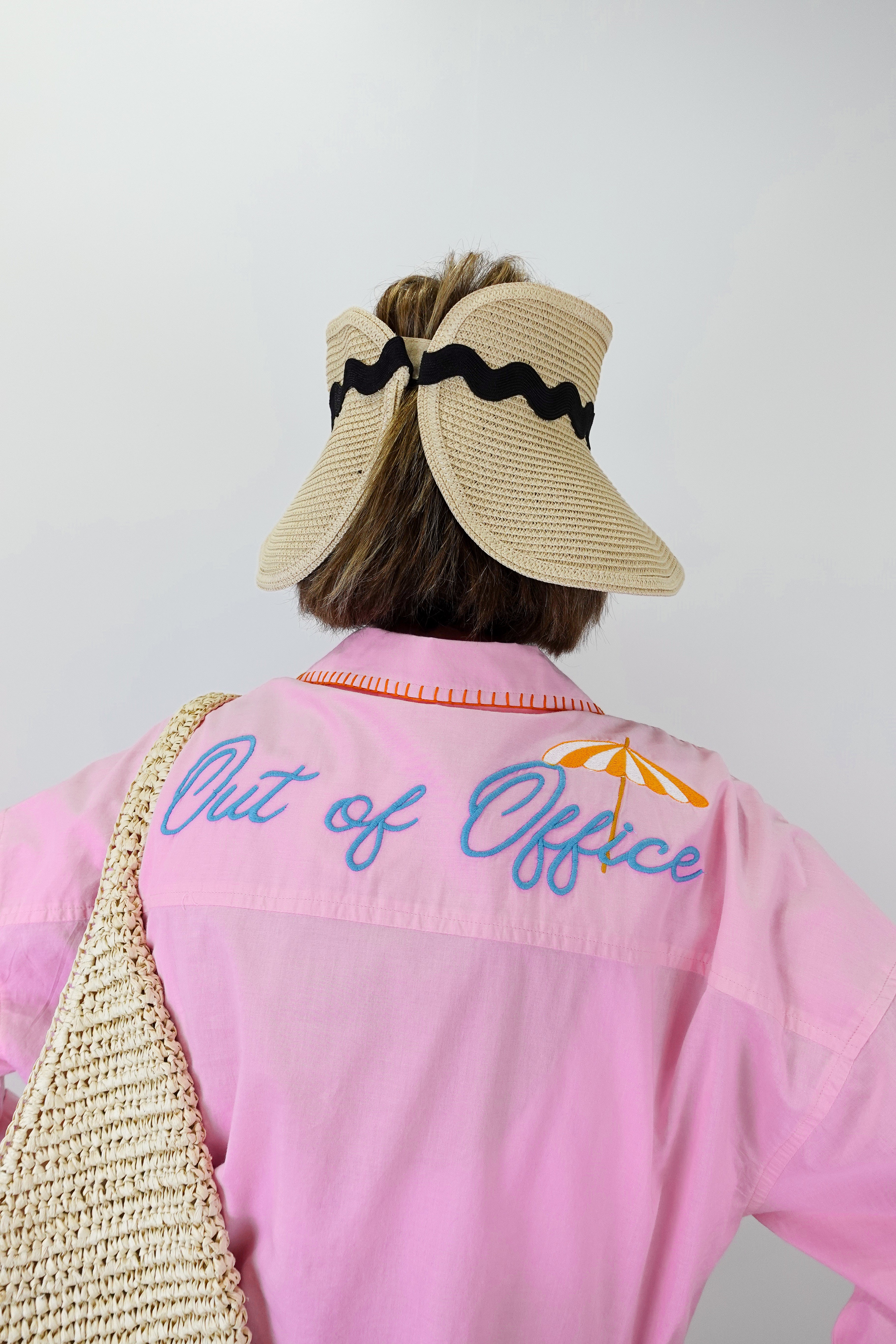 Person wearing a pink shirt with 'Out of Office' text, beige visor, and straw bag on a white background