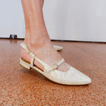 White pointed-toe shoes with gold straps on a brown floor.