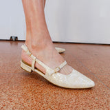 White pointed-toe shoes with gold straps on a brown floor.