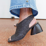 A close-up of a person wearing a black woven slingback mule sandal with a chunky block heel, paired with blue jeans, standing on a terrazzo floor.