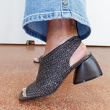 A close-up of a person wearing a black woven slingback mule sandal with a chunky block heel, paired with blue jeans, standing on a terrazzo floor.