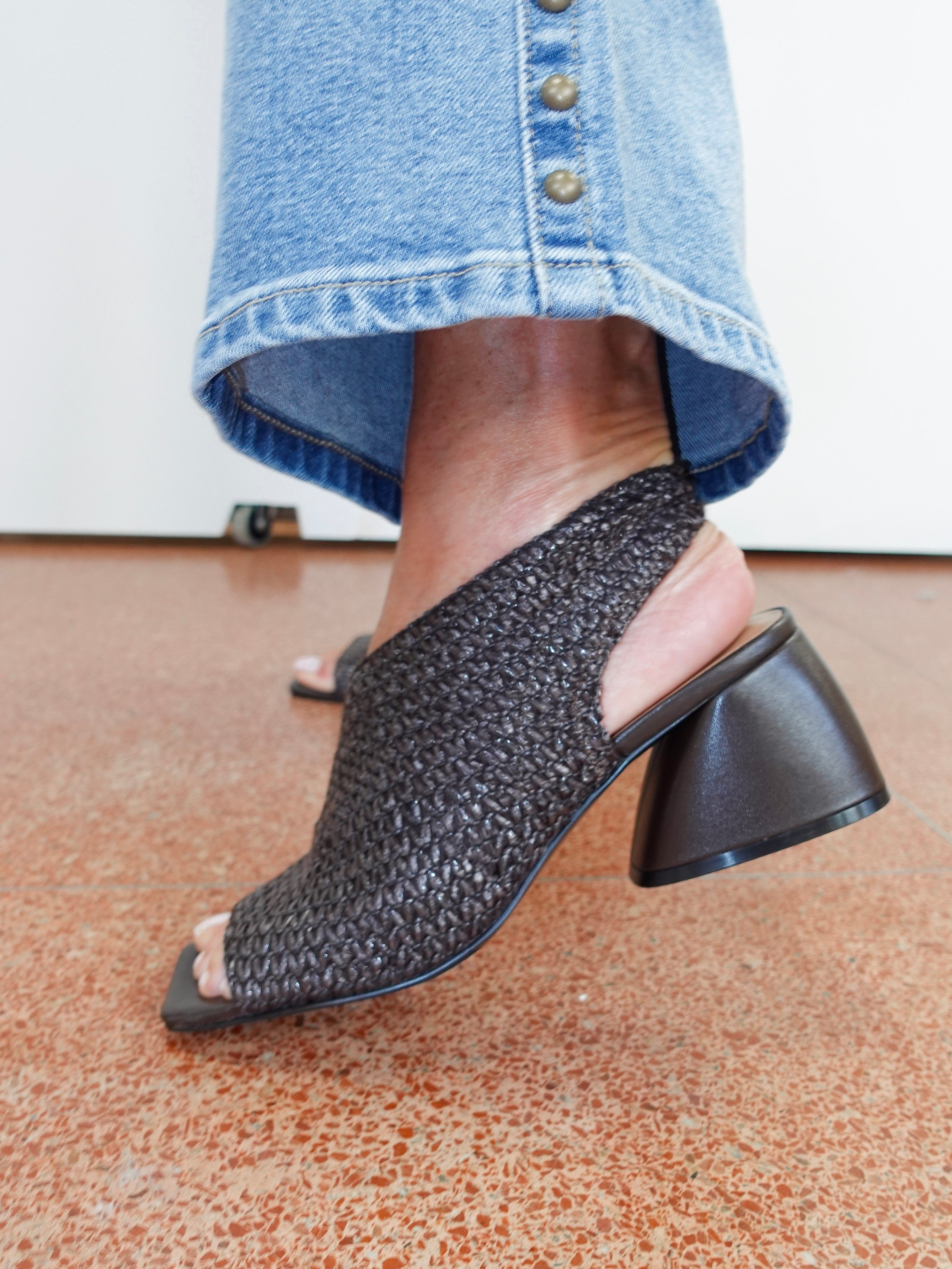 A close-up of a person wearing a black woven slingback mule sandal with a chunky block heel, paired with blue jeans, standing on a terrazzo floor.
