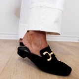 Black loafers with gold buckles worn by a person on a wooden floor.