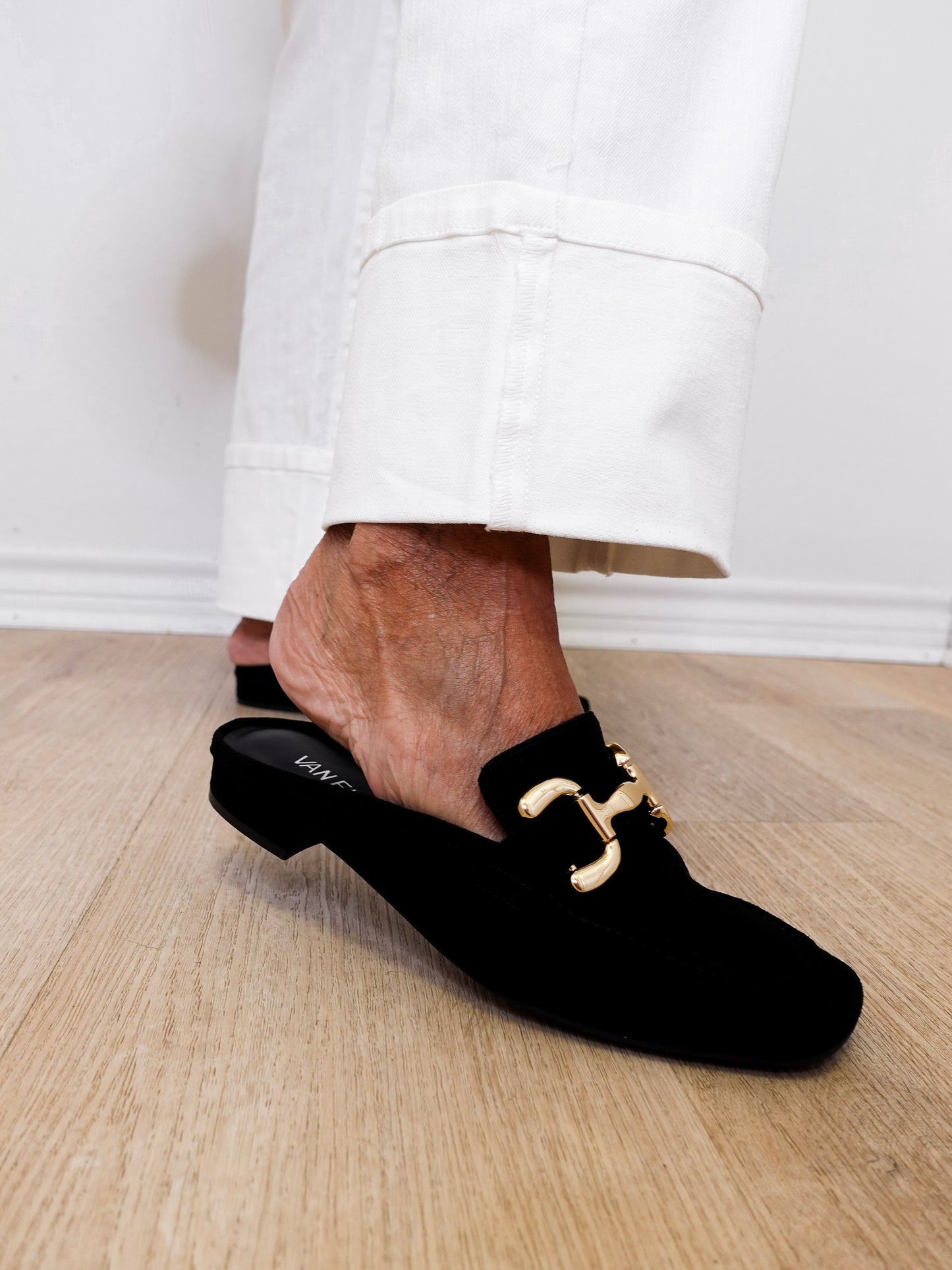 Black loafers with gold buckles worn by a person on a wooden floor.