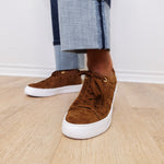 Brown suede sneakers with white soles worn with rolled-up jeans on a wooden floor.