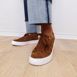 Brown suede sneakers with white soles worn with rolled-up jeans on a wooden floor.