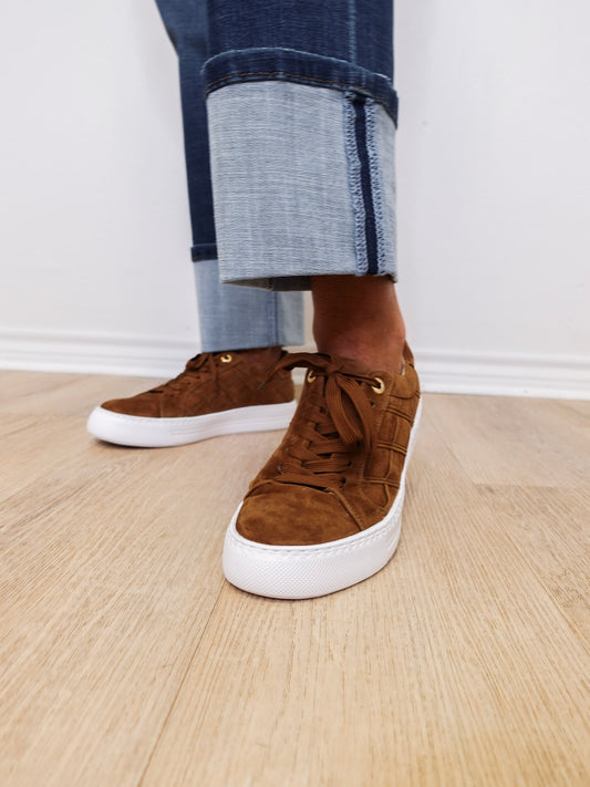 Brown suede sneakers with white soles worn with rolled-up jeans on a wooden floor.