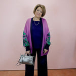 Woman wearing a purple cardigan with green patterns, holding a silver handbag against a pink wall.