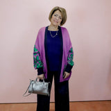 Woman wearing a purple cardigan with green patterns, holding a silver handbag against a pink wall.