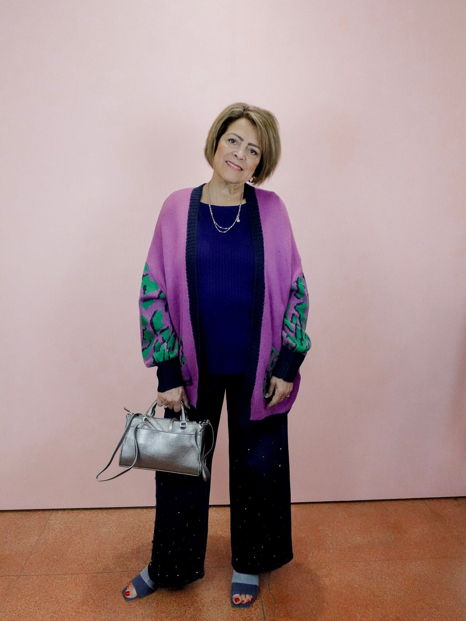 Woman wearing a purple cardigan with green patterns, holding a silver handbag against a pink wall.