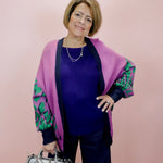 Woman wearing a purple cardigan with green patterns, holding a silver handbag against a pink background
