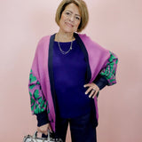 Woman wearing a purple cardigan with green patterns, holding a silver handbag against a pink background