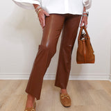 Person wearing brown leather pants and loafers, holding a brown handbag against a white wall.