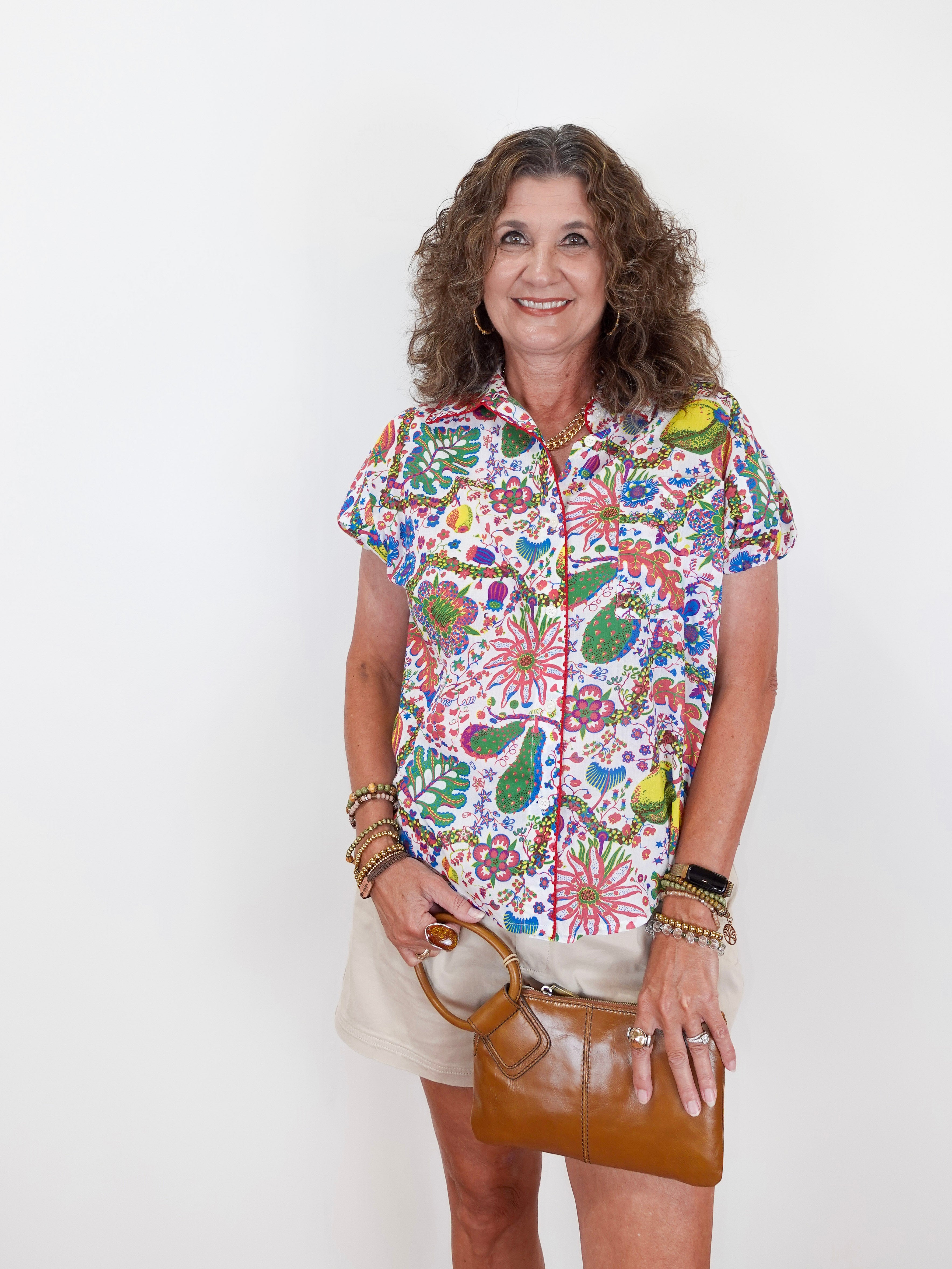 Woman wearing a colorful patterned shirt holding a brown handbag against a white background