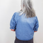A person viewed from behind wearing a light blue denim shirt with puffed three-quarter sleeves and dark gray pants, standing against a plain off-white wall.