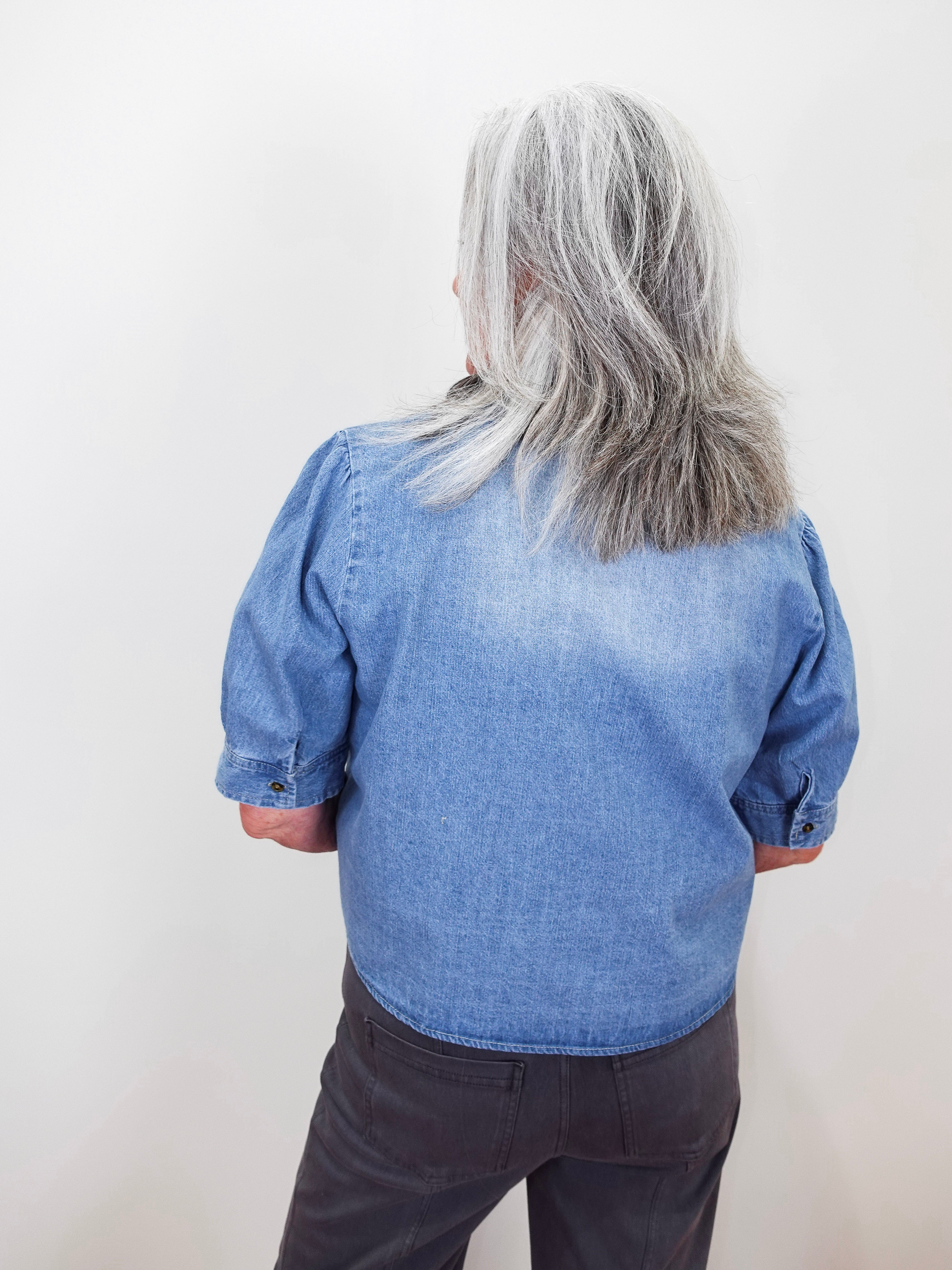 A person viewed from behind wearing a light blue denim shirt with puffed three-quarter sleeves and dark gray pants, standing against a plain off-white wall.
