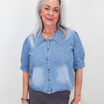 An older woman with gray hair wearing a light blue denim short-sleeve button-up shirt and dark gray pants, standing against a plain white background.