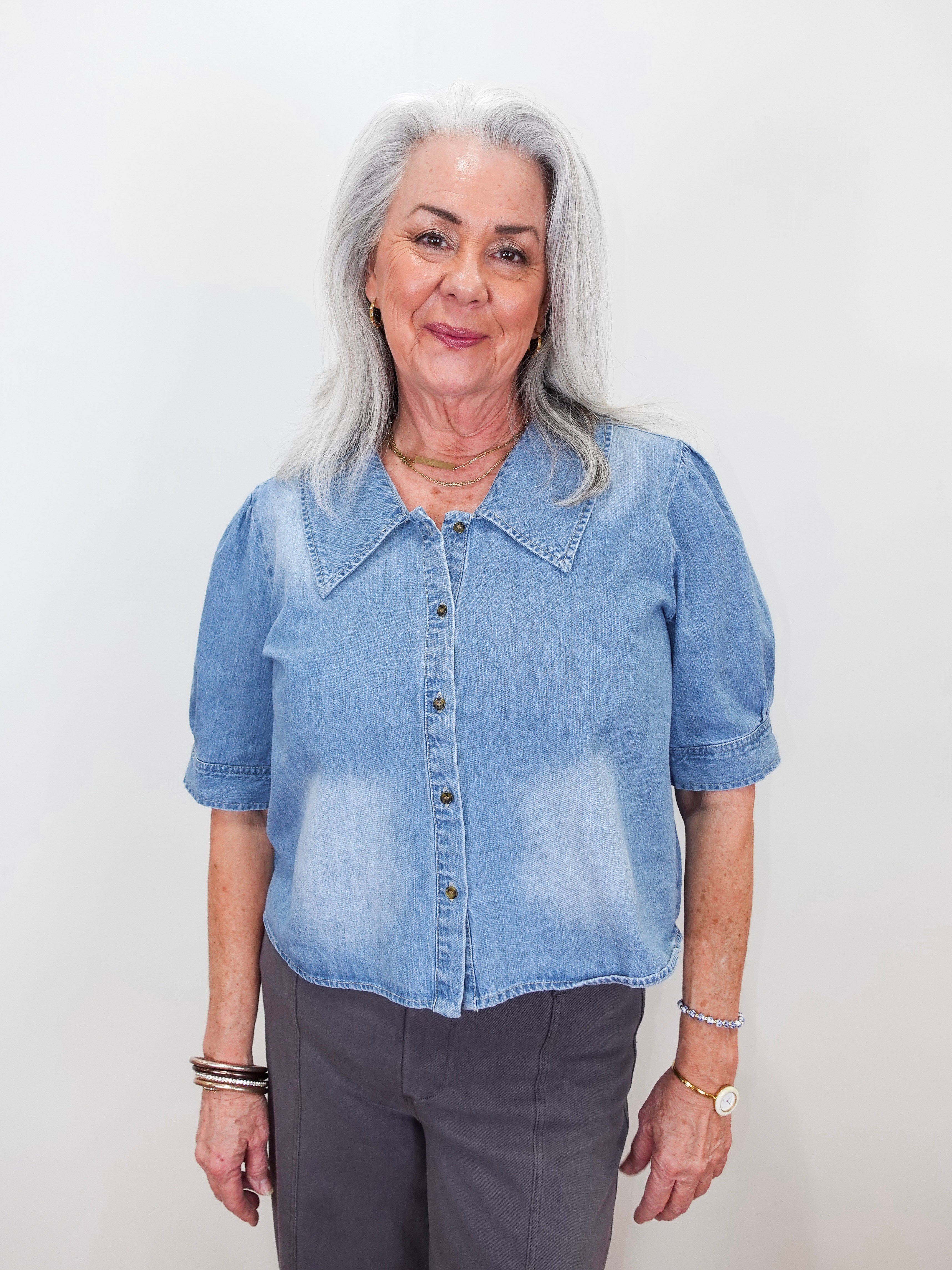 An older woman with gray hair wearing a light blue denim short-sleeve button-up shirt and dark gray pants, standing against a plain white background.