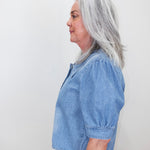 A woman with shoulder-length gray hair is shown in profile wearing a light blue cropped short-sleeve blouse with puff sleeves and dark gray pants, standing against a plain off-white wall.