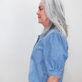 A woman with shoulder-length gray hair is shown in profile wearing a light blue cropped short-sleeve blouse with puff sleeves and dark gray pants, standing against a plain off-white wall.